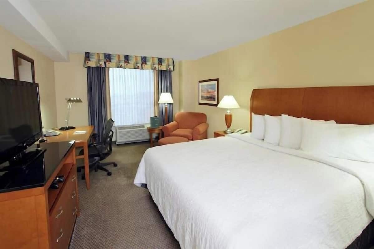 Une chambre d’hôtel avec un lit, un bureau avec une télévision, une chaise, une lampe et une fenêtre avec des rideaux.