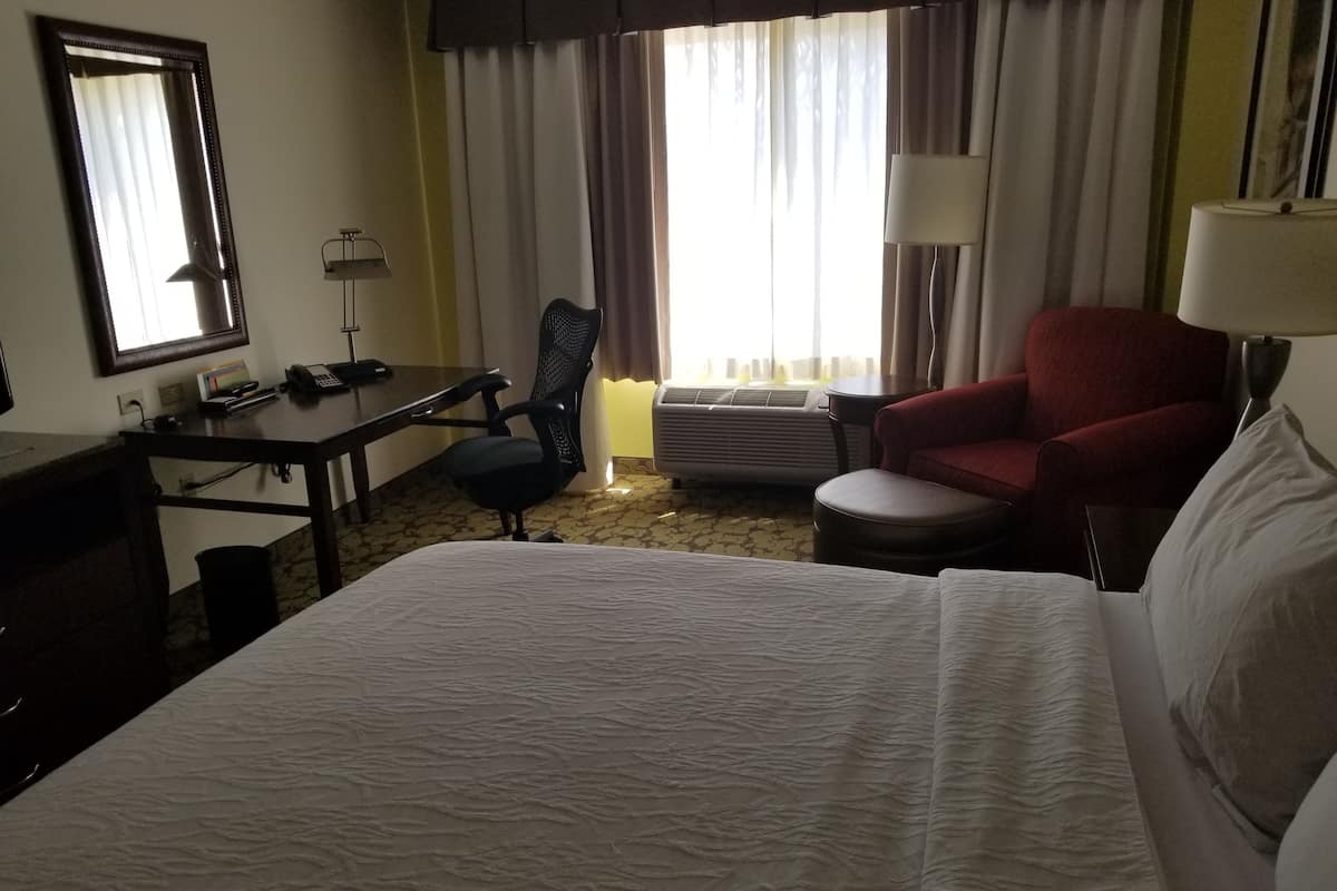Une chambre d’hôtel avec un lit, un bureau, une chaise, un fauteuil rouge et une lampe.
