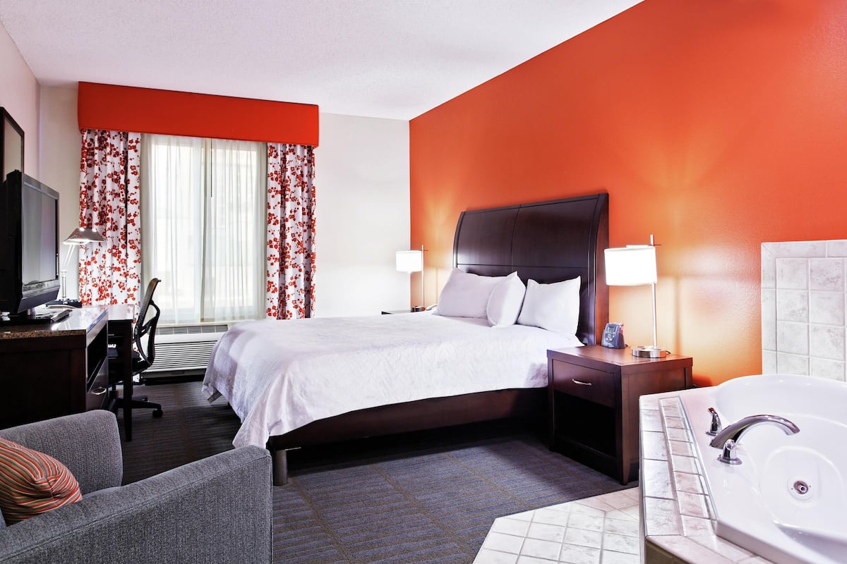Quarto de hotel com cama, escrivaninha com computador, cadeira, luminária de mesa, mesa de cabeceira com abajur e banheiro com banheira e pia.