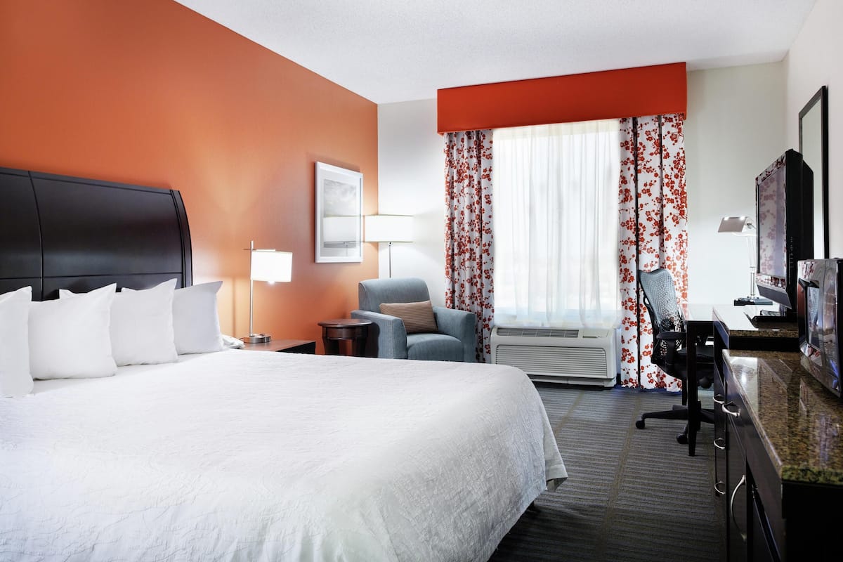 Quarto de hotel com uma cama grande, uma mesa de escritório com computador, uma cadeira, uma televisão e uma janela com cortinas.