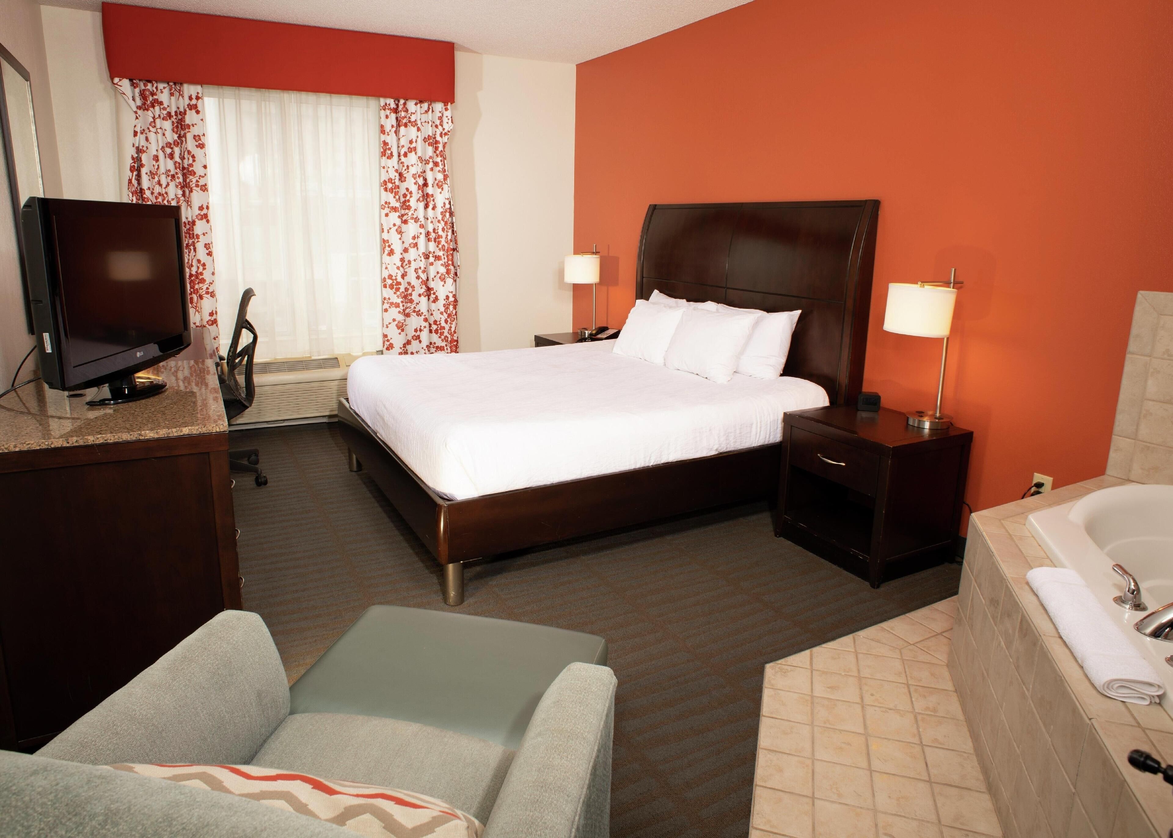 Une chambre d’hôtel comprenant un lit, un bureau avec un ordinateur, une chaise, une table de chevet avec une lampe, et une salle de bain avec une baignoire.