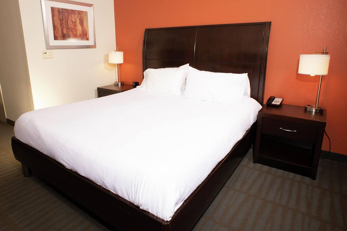 Quarto de hotel com uma cama grande, duas mesas de cabeceira com abajures, um telefone fixado na parede e um quadro emoldurado.
