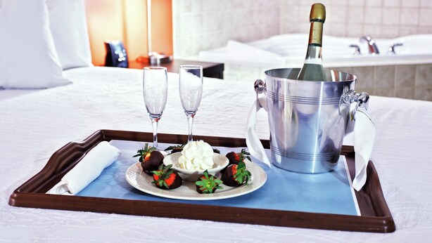 A tray with a bottle of champagne, two glasses, a bowl of whipped cream, and chocolate strawberries on a bed.