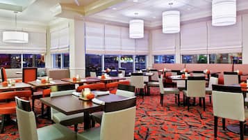 A hotel dining area with tables and chairs, large windows, and hanging lights.