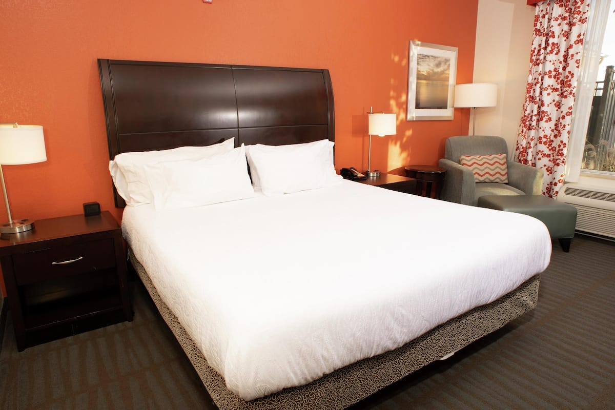 Quarto de hotel com uma cama grande, duas mesinhas de cabeceira, uma cadeira, um abajur e um quadro na parede.