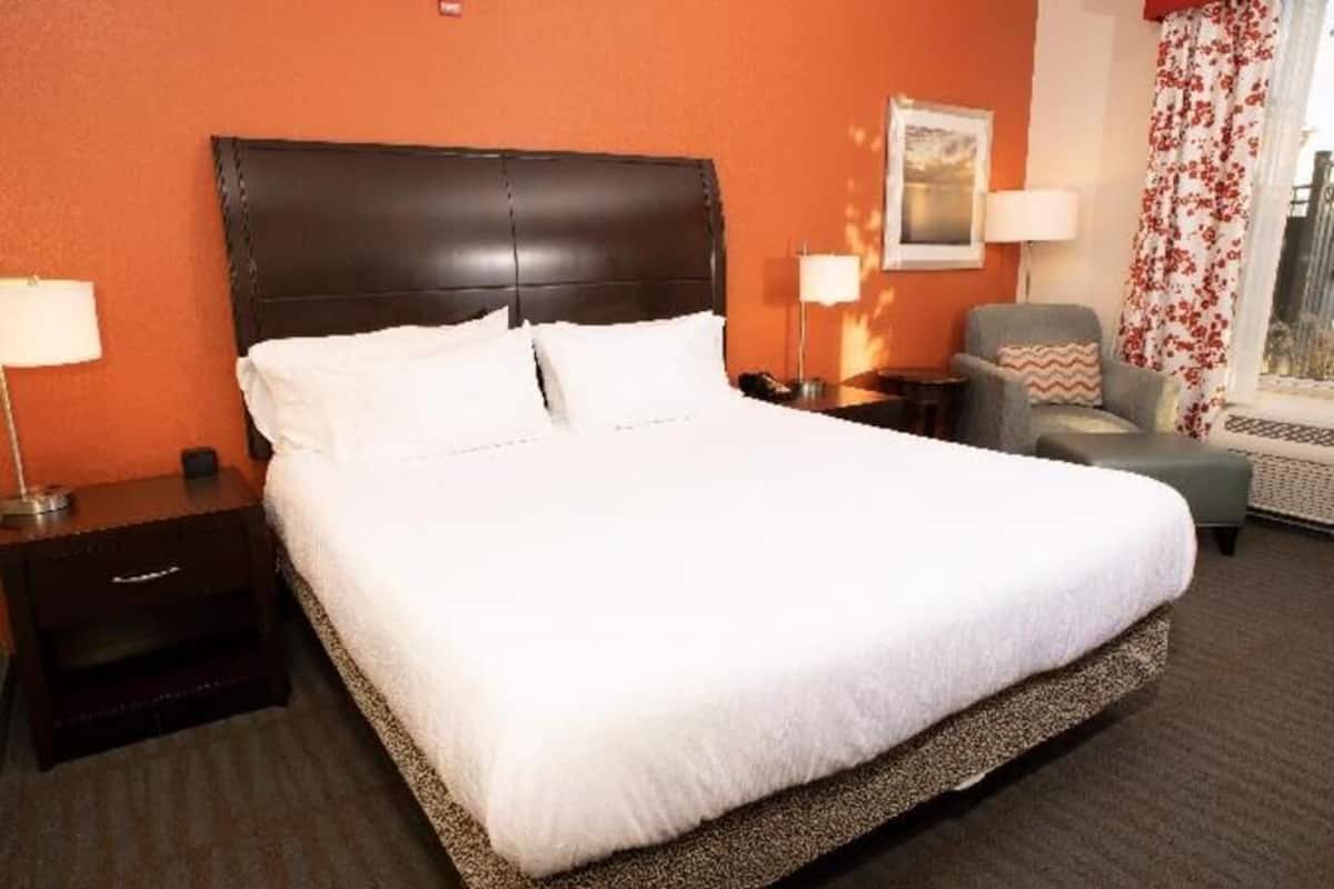 Quarto de hotel com uma cama grande, uma mesa de cabeceira, uma cadeira e uma janela com cortinas.