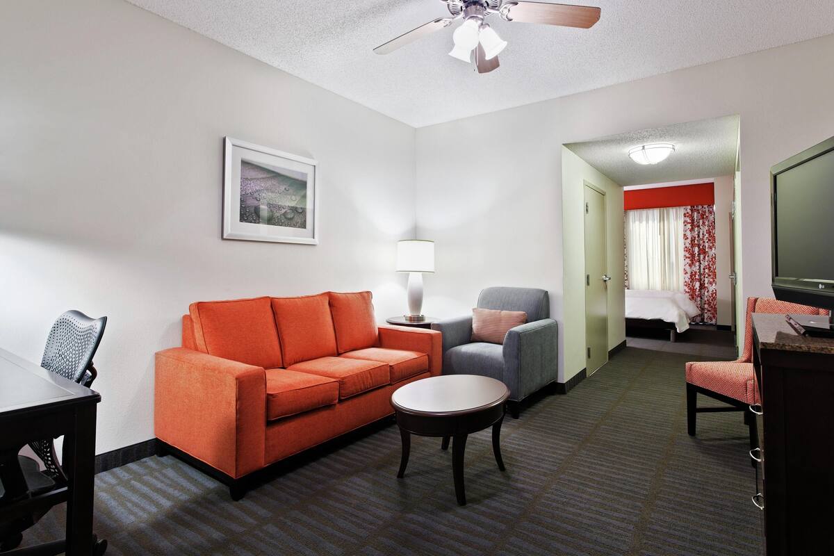 Quarto de hotel com sofá, poltrona, mesa pequena, abajur, cama e televisão.