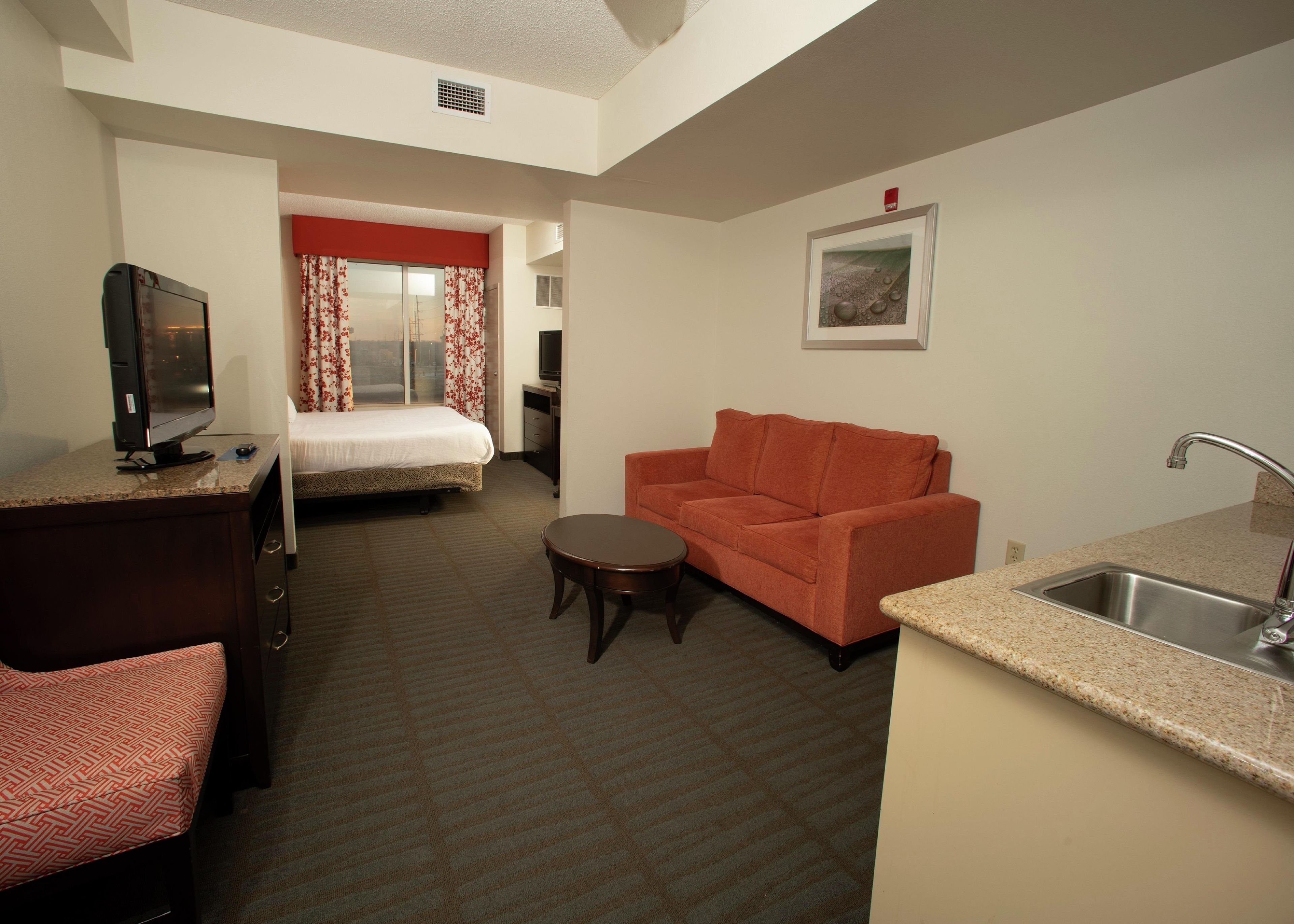 Habitación de hotel con cama, televisión, sofá, una mesita y una zona de cocina con lavabo.