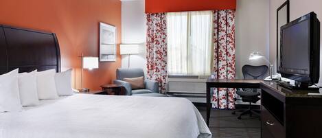 Une chambre d’hôtel avec un grand lit, un bureau avec un ordinateur, une télévision et une fenêtre agrémentée de rideaux à motifs floraux.