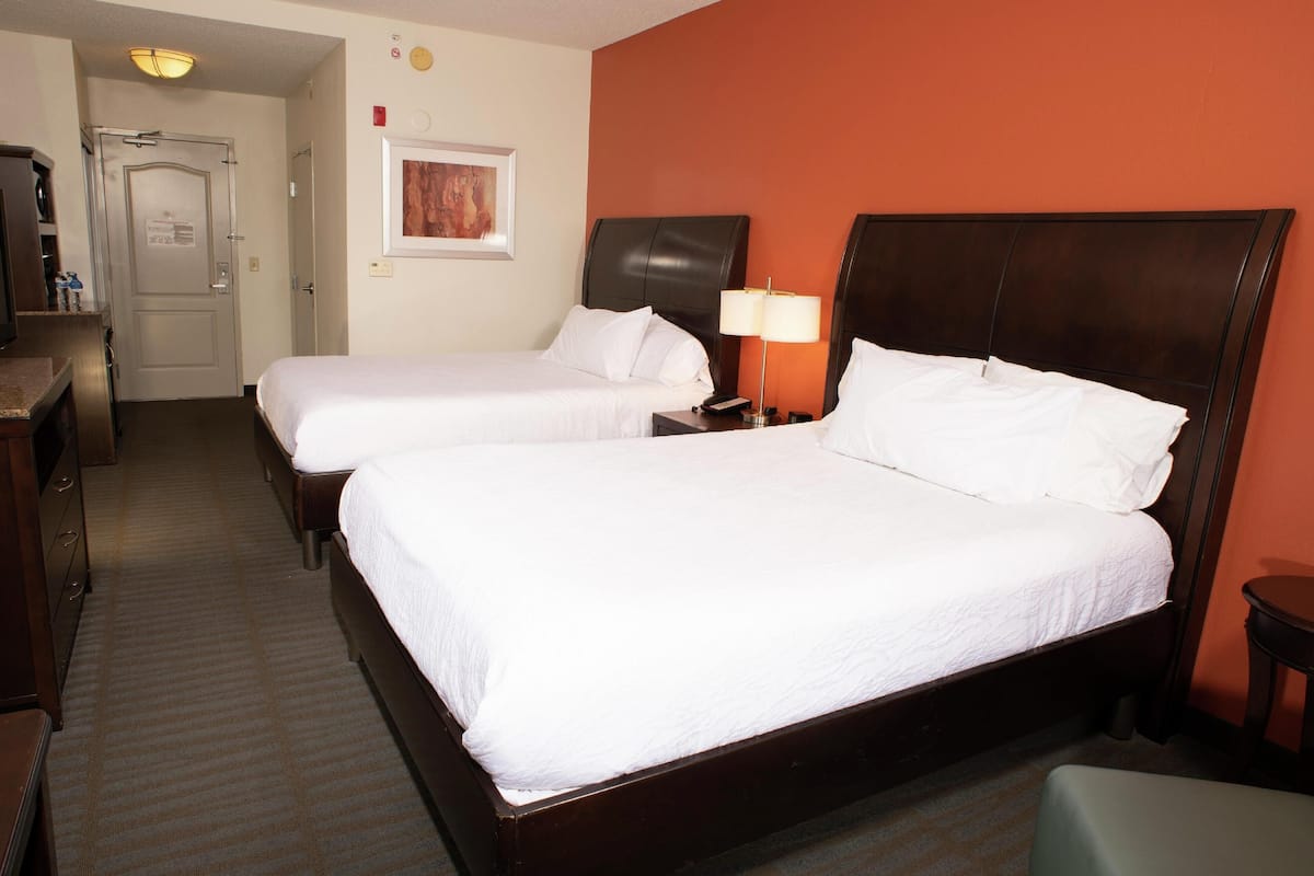 Quarto de hotel com duas camas de solteiro, uma escrivaninha, uma cadeira e um abajur.