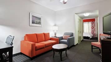 A hotel room with a sofa, armchair, small table, lamp, bed, and a television.