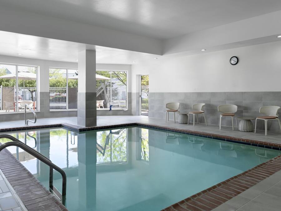 Un área de piscina interior moderna con paredes de vidrio transparente, sillas blancas y un reloj en la pared.