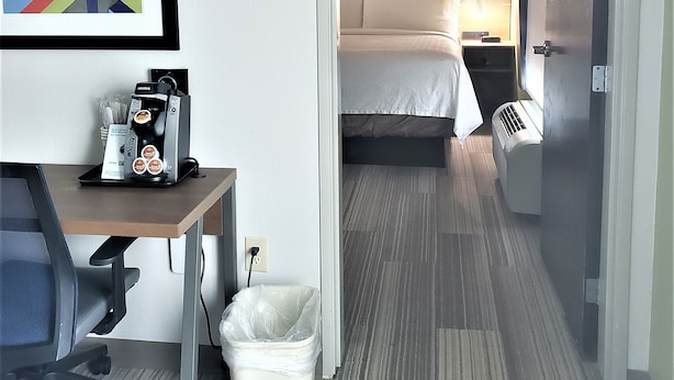 Habitación de hotel con cama, mesita de noche y escritorio con cafetera. Hay un cubo de basura y un cuadro en la pared.