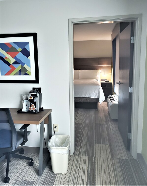 Habitación de hotel con cama, mesita de noche y escritorio con cafetera. Hay un cubo de basura y un cuadro en la pared.