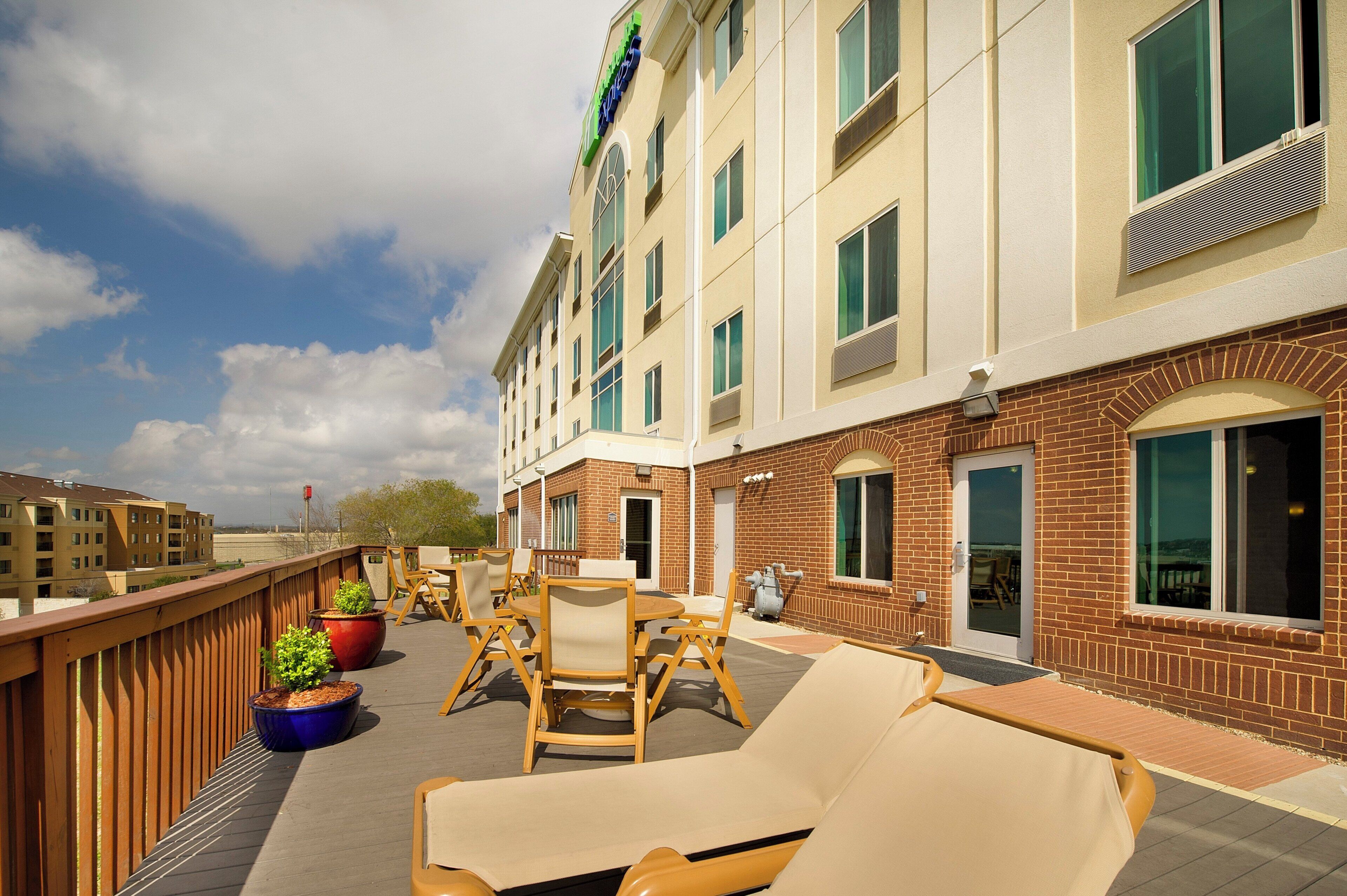 A hotel exterior with a covered outdoor seating area, including lounge chairs and tables, and a brick building with multiple windows.