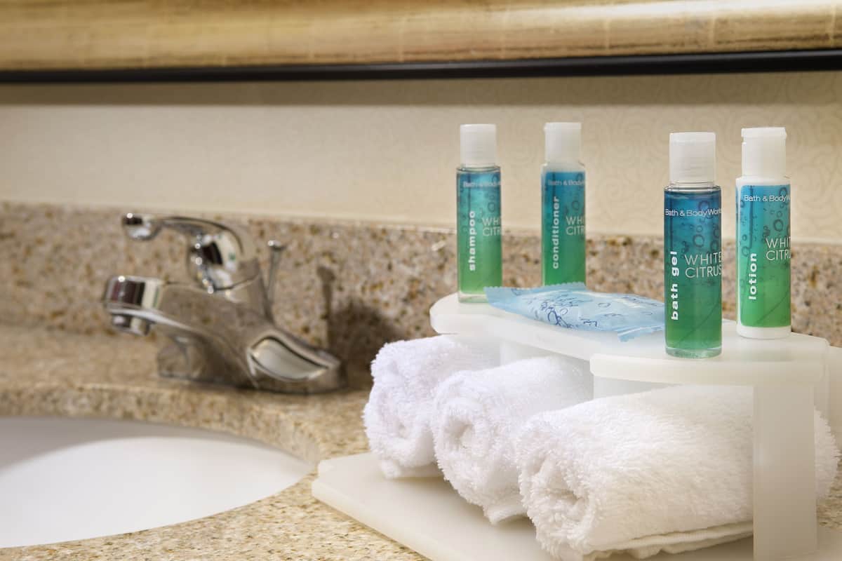 A bathroom sink with a granite countertop, a white towel rack holding two white towels, and three bottles of white citrus shampoo and conditioner.