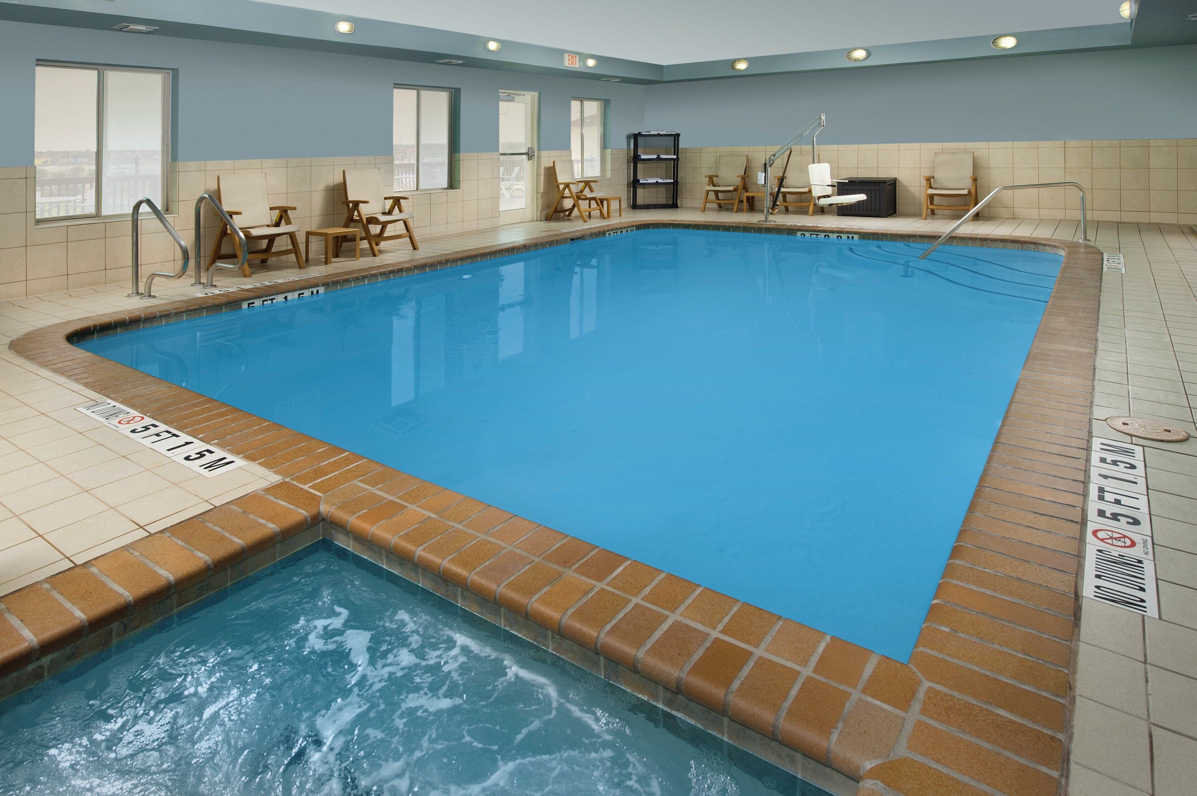 A rectangular indoor swimming pool with a tiled edge, a shallow end marked with depth indicators, and multiple wooden chairs and tables around the perimeter.