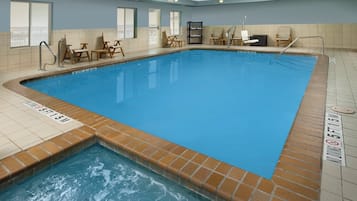 A rectangular indoor swimming pool with a tiled edge, a shallow end marked with depth indicators, and multiple wooden chairs and tables around the perimeter.