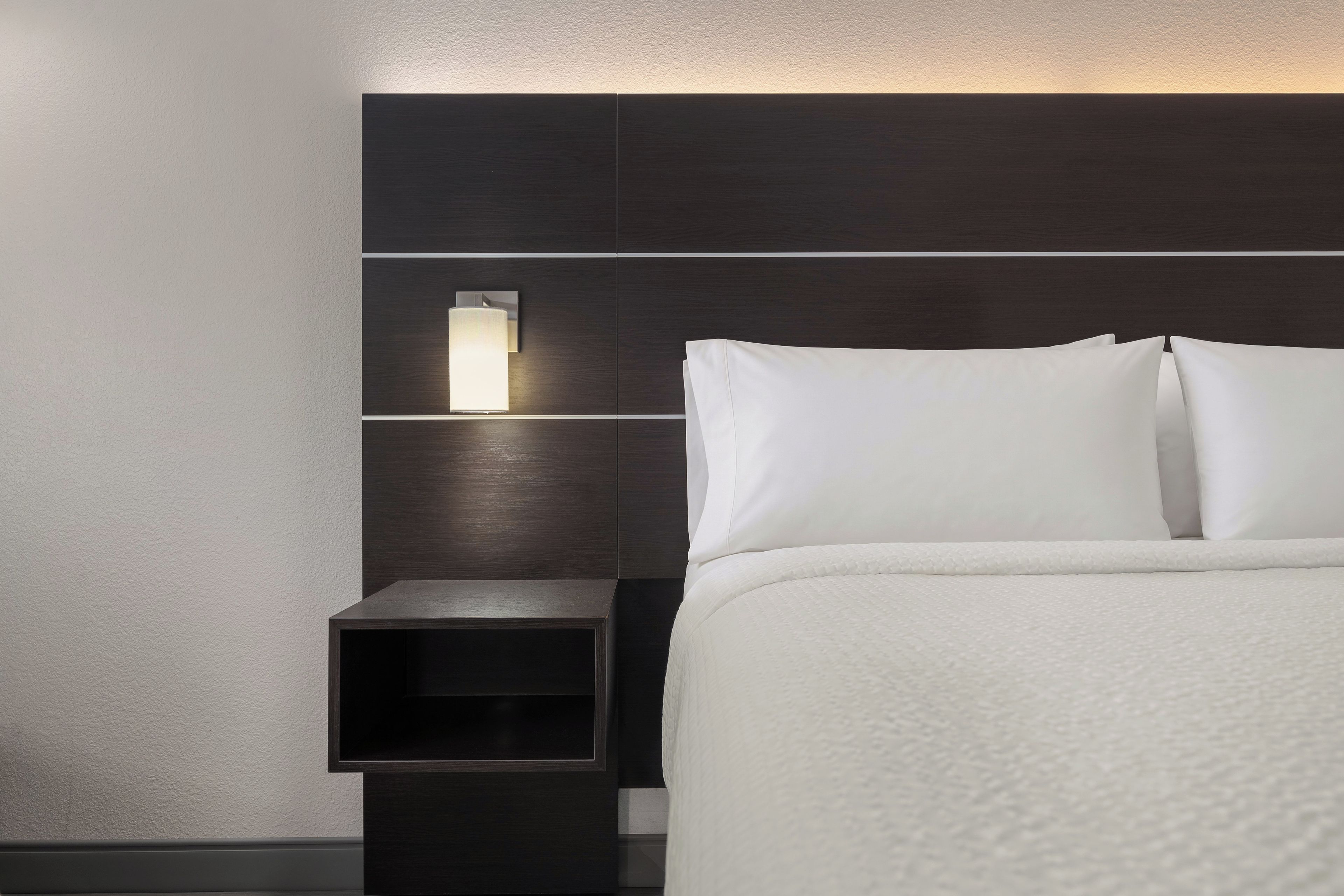 A hotel room with a bed, a nightstand, and a wall-mounted light fixture.