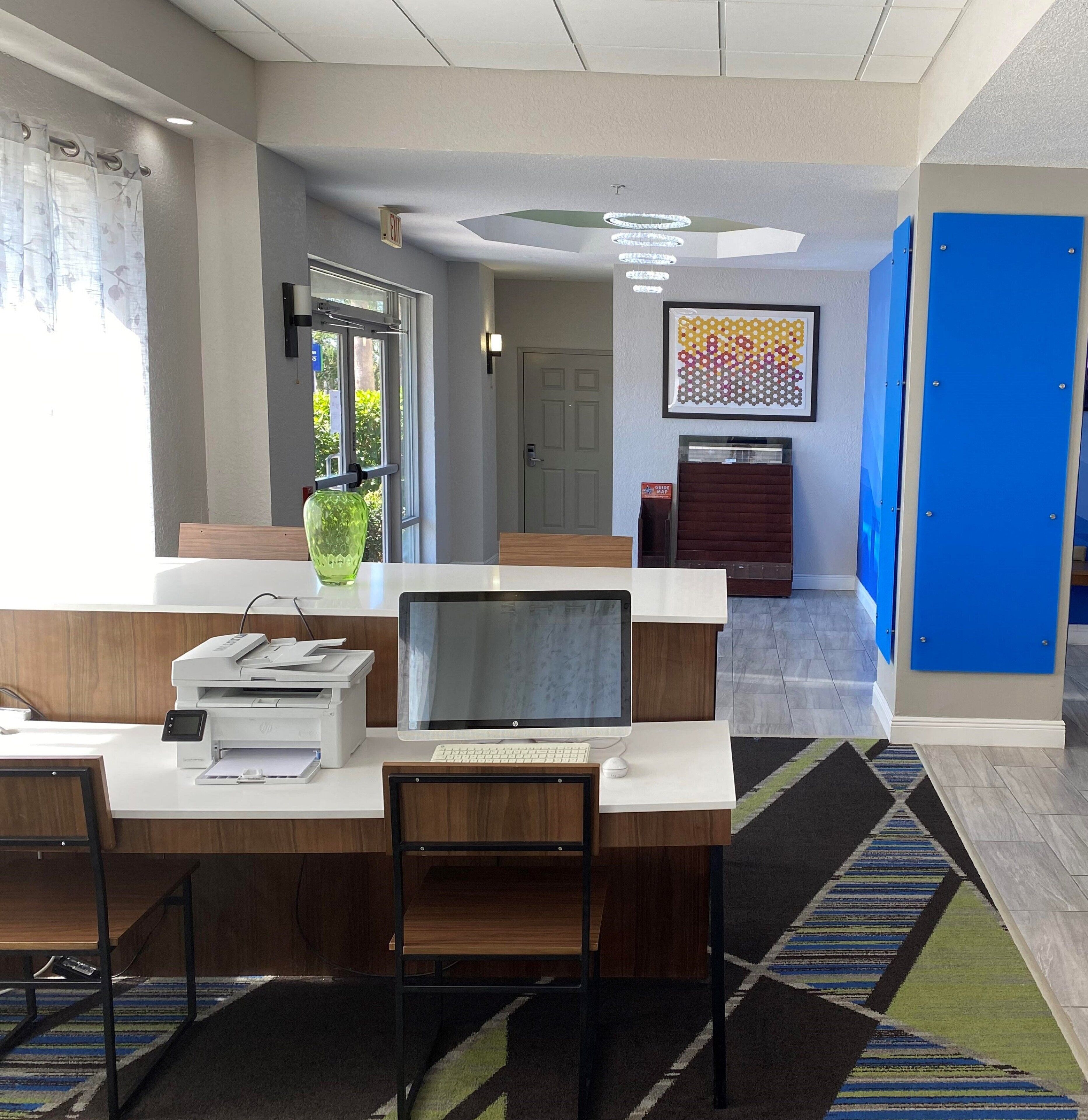 A modern reception desk with a computer, printer, and seating area. There is a door with a sign, a framed artwork, and a blue cabinet.