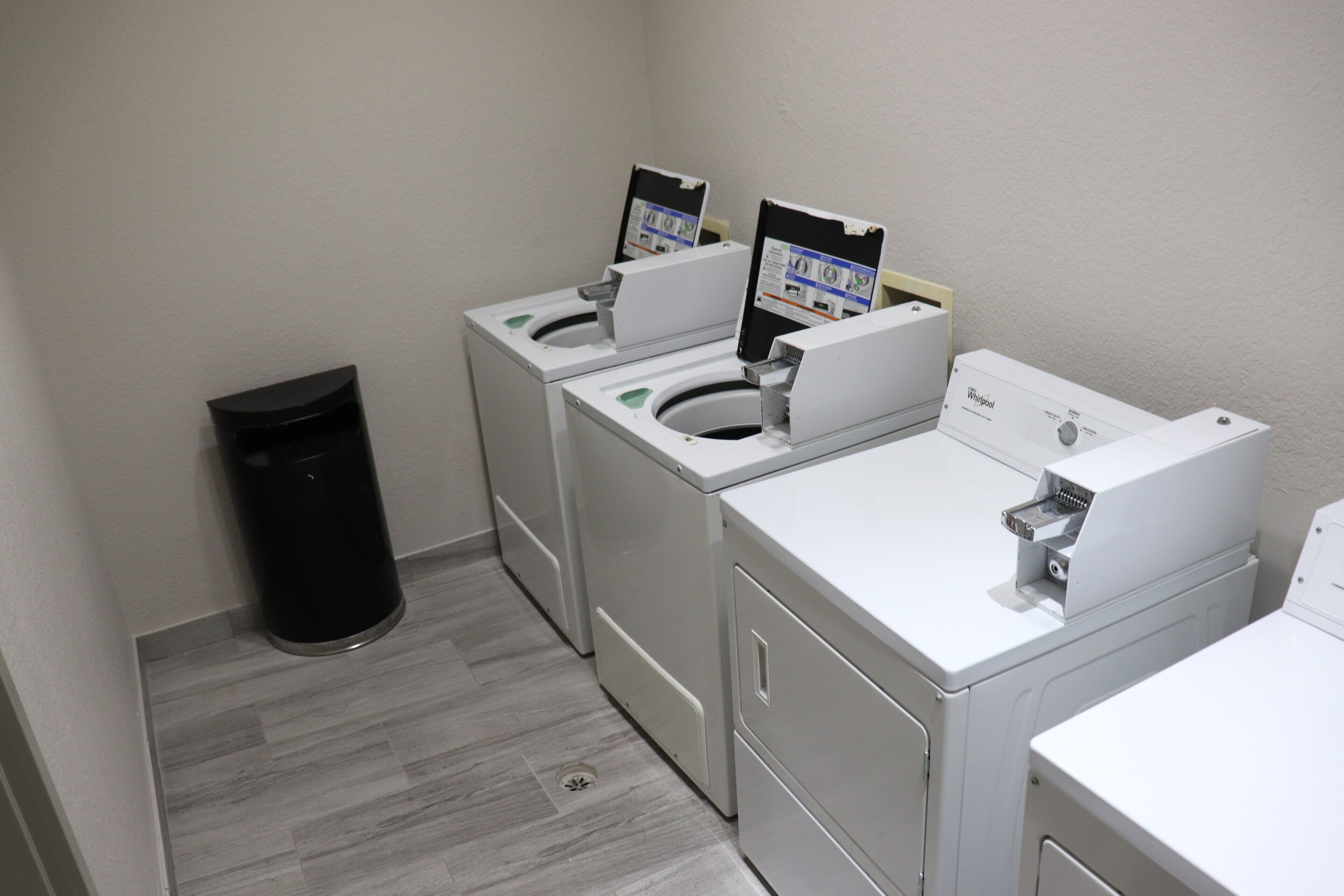 A laundry room with two front-loading washers and dryers, a trash can, and a floor drain.