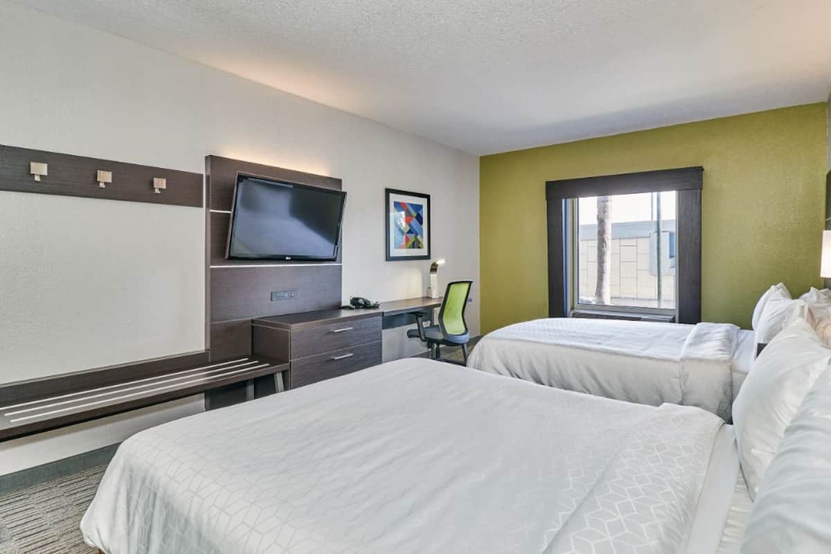 A hotel room with two beds, a desk with a chair, a flat-screen TV, and a window with blinds.