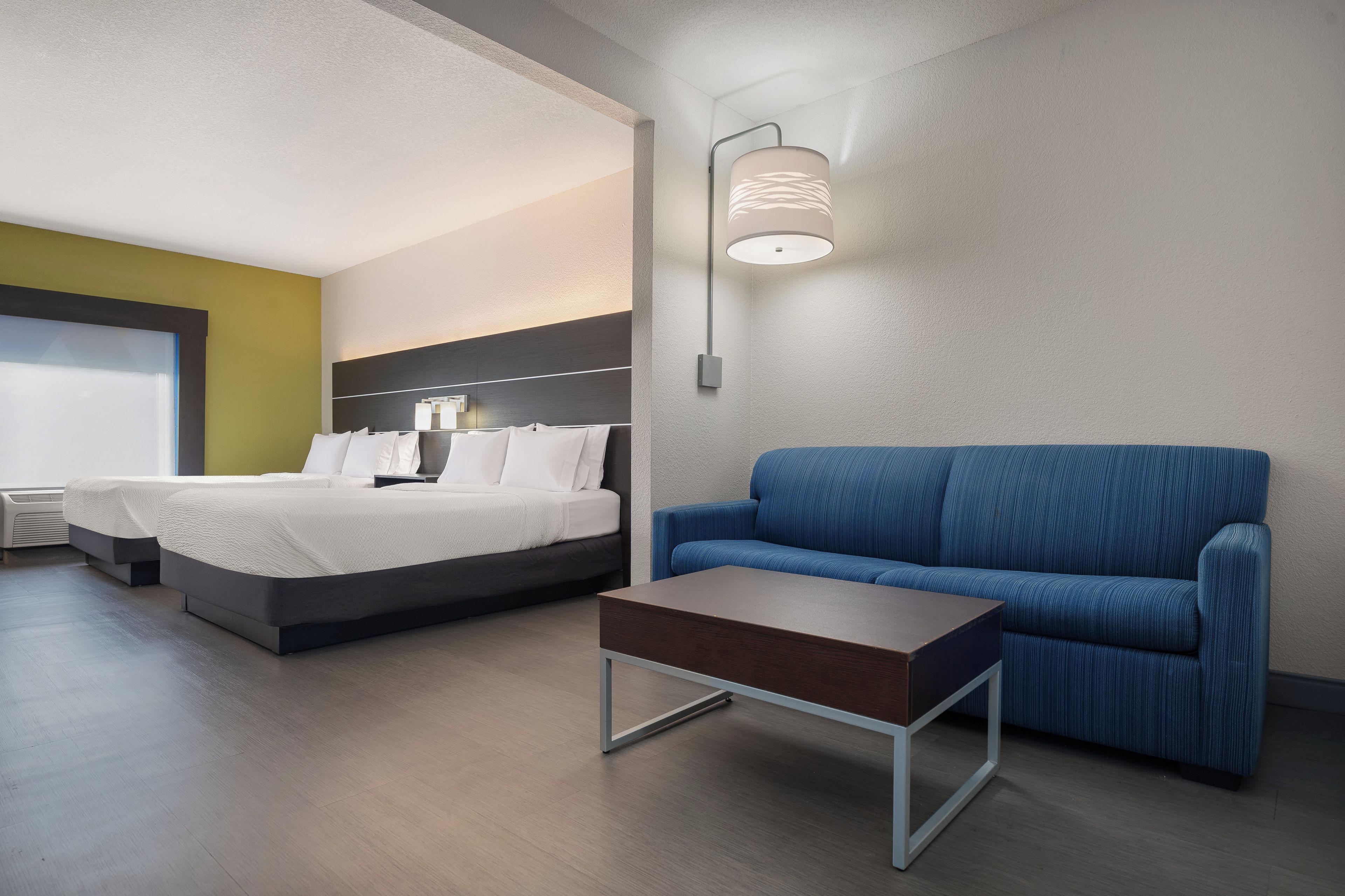 A modern hotel room with a bed, a sofa, a small table, and a wall-mounted lamp.