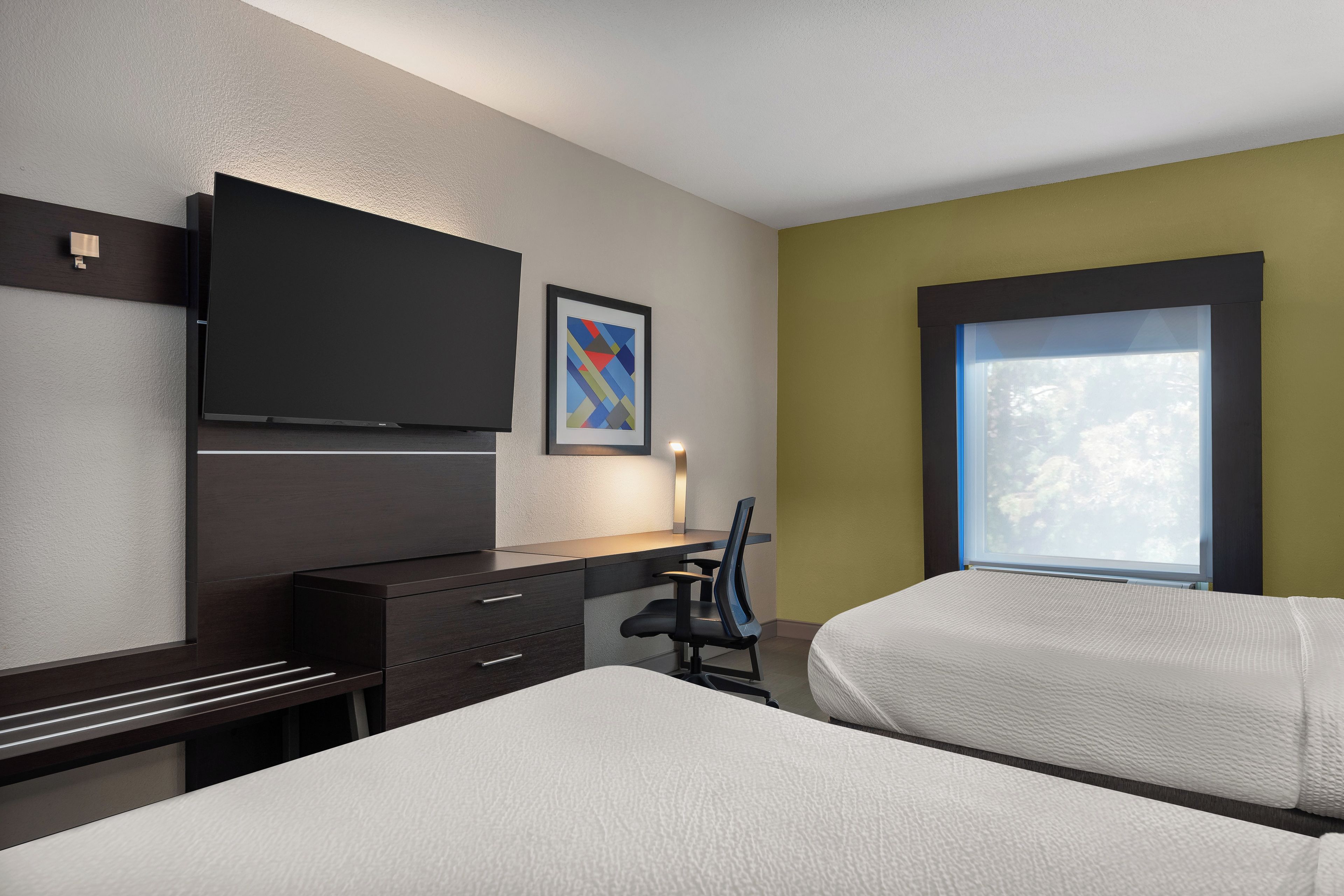 A hotel room with a bed, a desk with drawers, a chair, a flat-screen TV mounted on the wall, a framed picture, and a window with a view of trees.