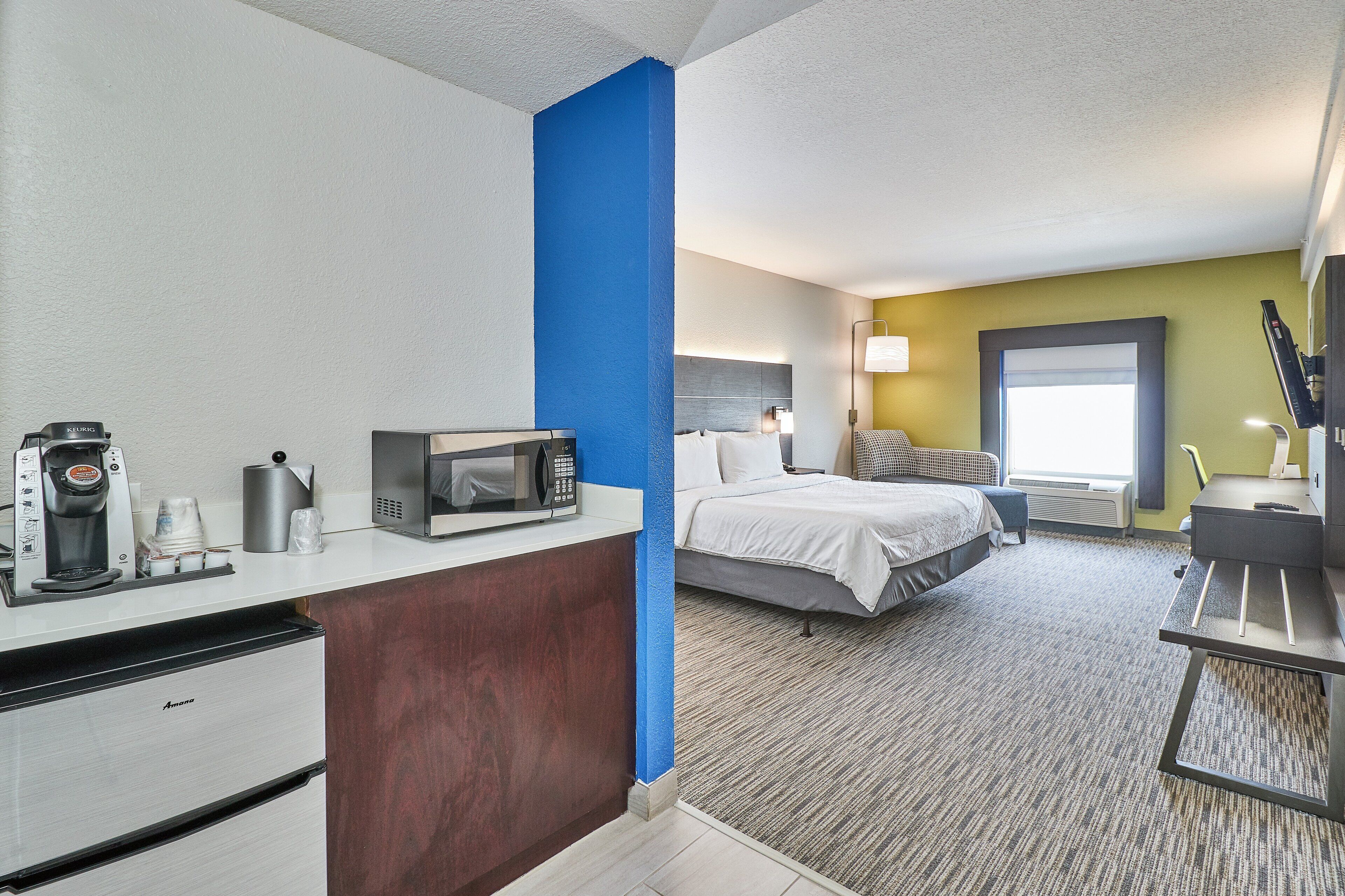 A hotel room with a kitchenette, a bed, a desk, and a window.