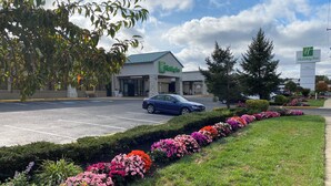 A Holiday Inn hotel with a parking lot, a blue car parked in front, and a flower bed with pink and orange flowers. - Holiday Inn Hazlet by IHG (Hazlet)