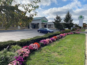 A Holiday Inn hotel with a parking lot, a blue car parked in front, and a flower bed with pink and orange flowers. - Holiday Inn Hazlet by IHG (Hazlet)
