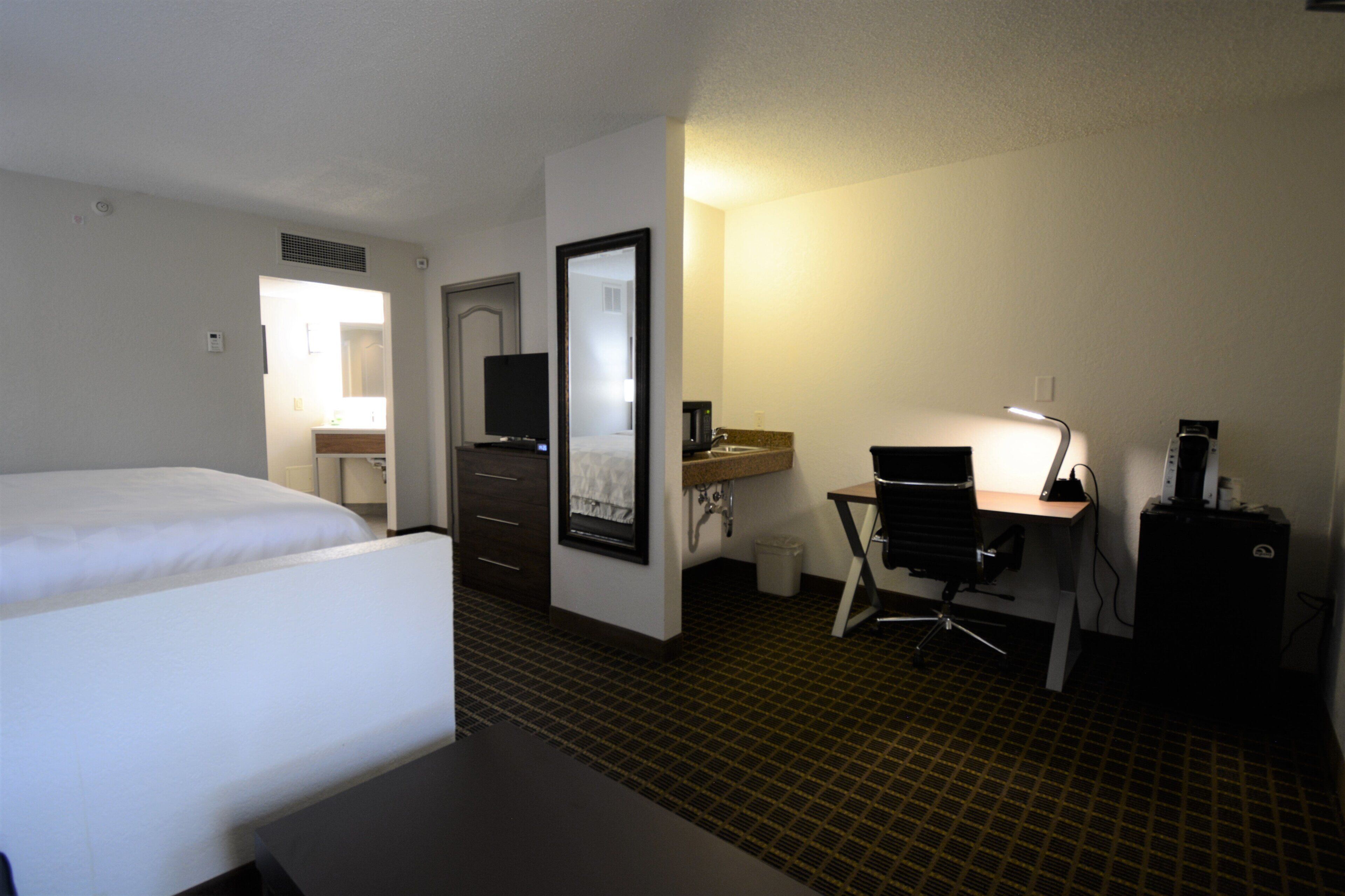 A hotel room with a bed, a desk with a chair, a mirror, a television, and a small kitchenette.