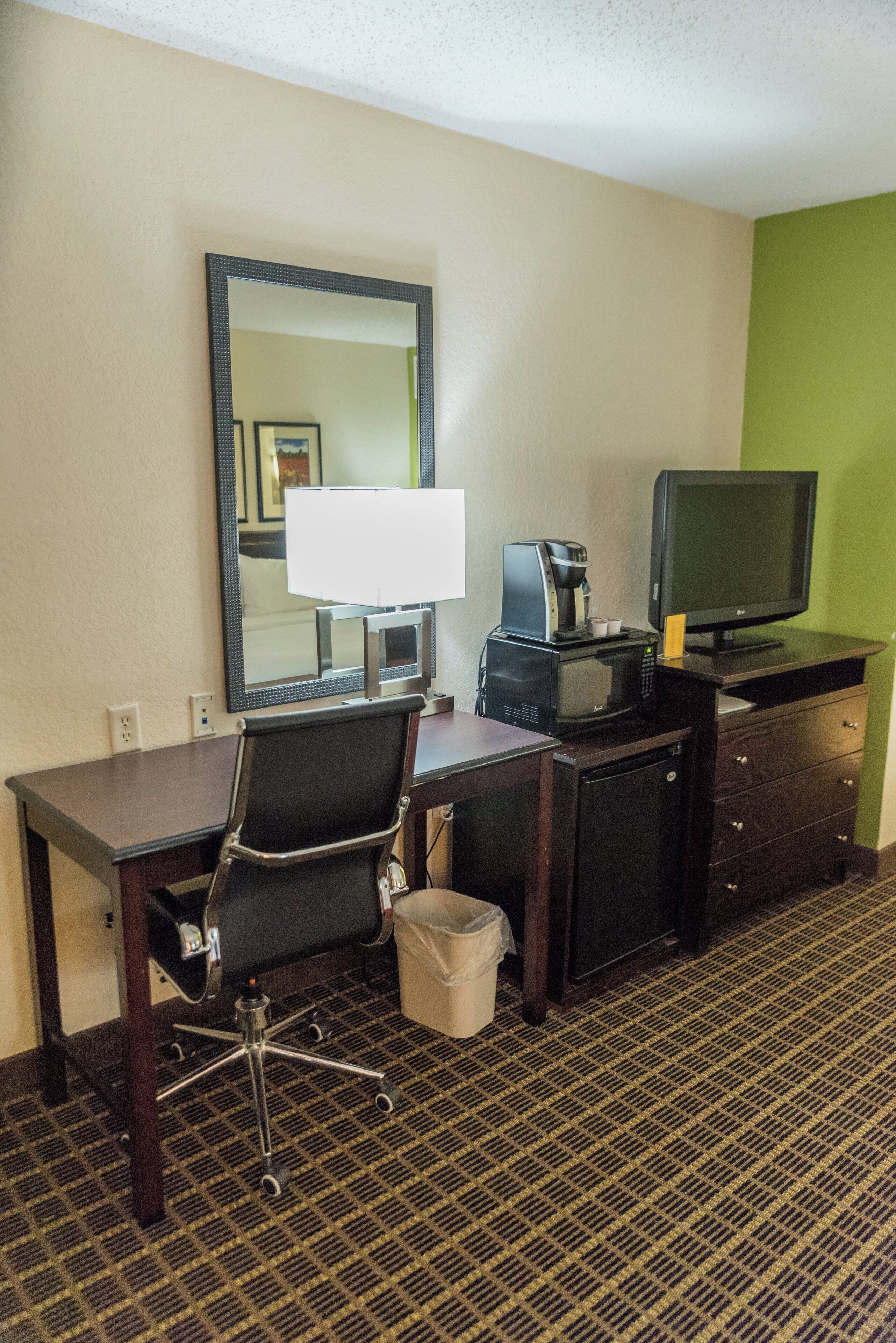 A hotel room with a desk, chair, television, coffee maker, and a trash bin.