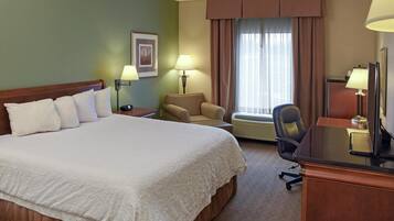 Ein Hotelzimmer mit einem großen Bett, einem Schreibtisch mit Computer, einem Sessel und einem Fenster mit Vorhängen.