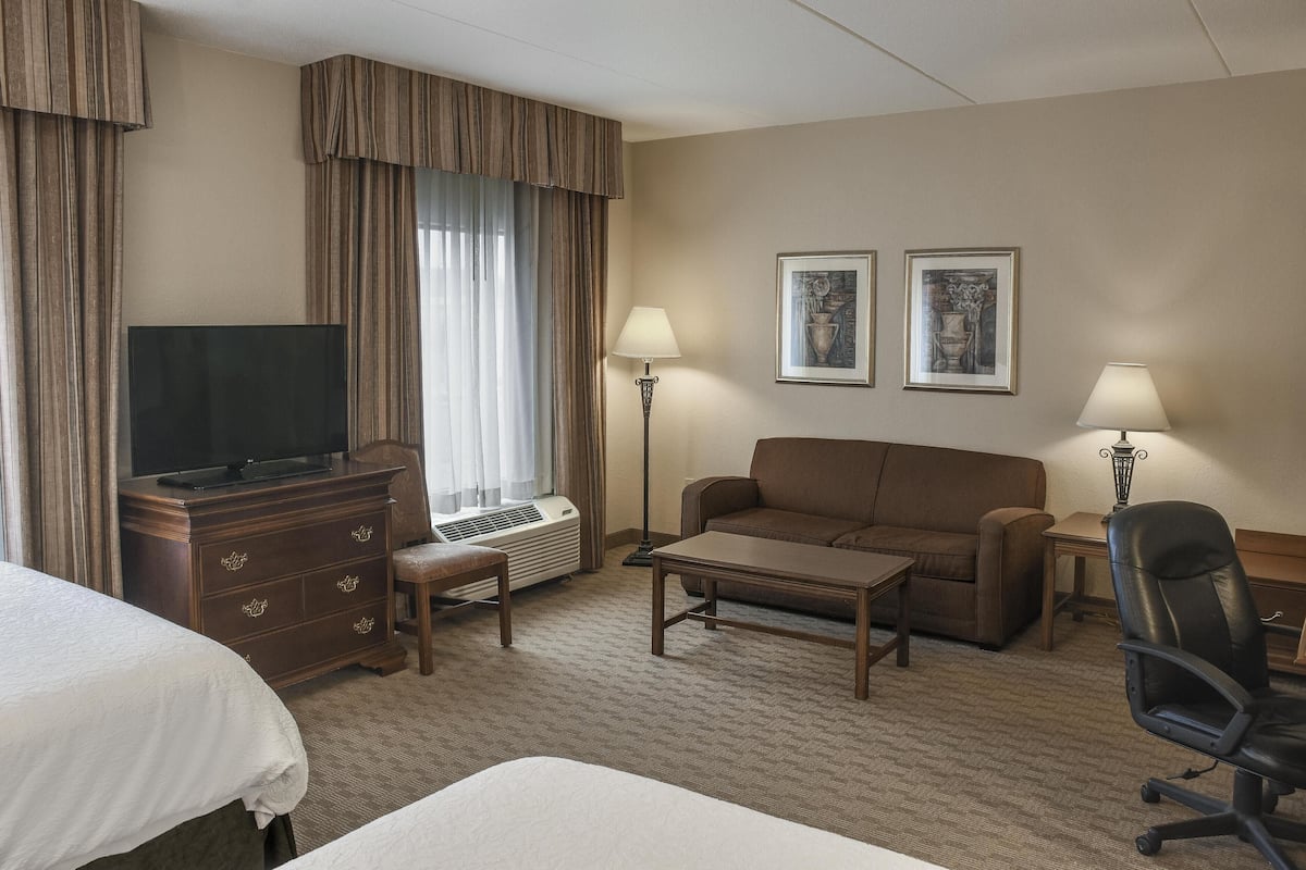 A hotel room with a flat-screen TV on a wooden dresser, a sofa, a coffee table, a desk with a lamp, and two framed pictures on the wall.