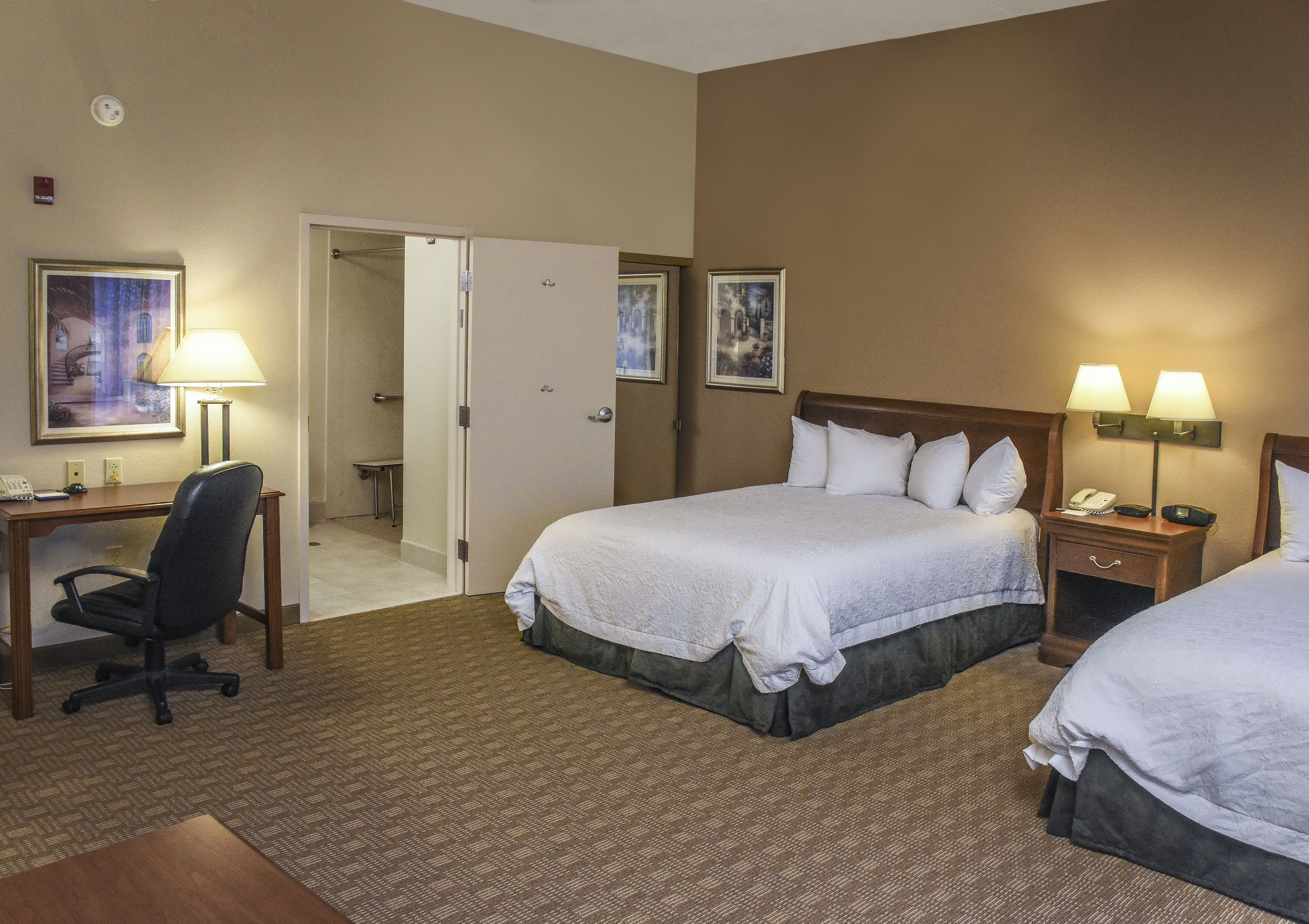 Une chambre d’hôtel avec deux lits, un bureau et une chaise. On y trouve deux tables de chevet avec des lampes, une porte donnant sur une salle de bain, et une petite table avec une chaise.