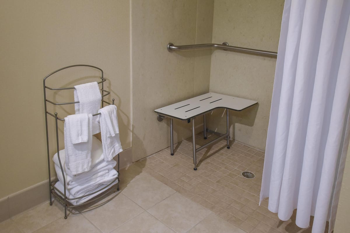 A bathroom with a metal towel rack holding folded towels, a small table, and a shower area with a grab bar.