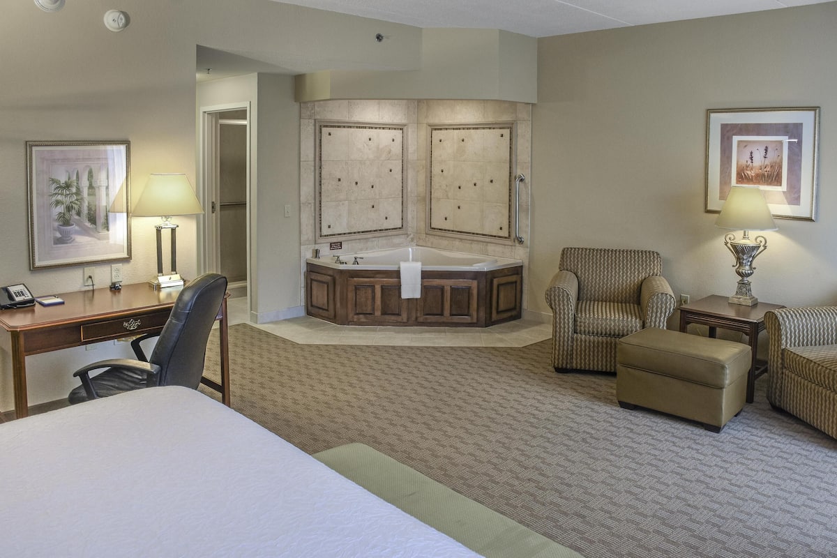A hotel room with a large bed, a desk with a chair, a sitting area with a chair and ottoman, a bathroom with a bathtub, and two framed pictures on the walls.