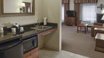 Quarto de hotel com cozinha compacta, incluindo micro-ondas, pia e armários. Há uma televisão, uma pequena mesa e um sofá na sala de estar.