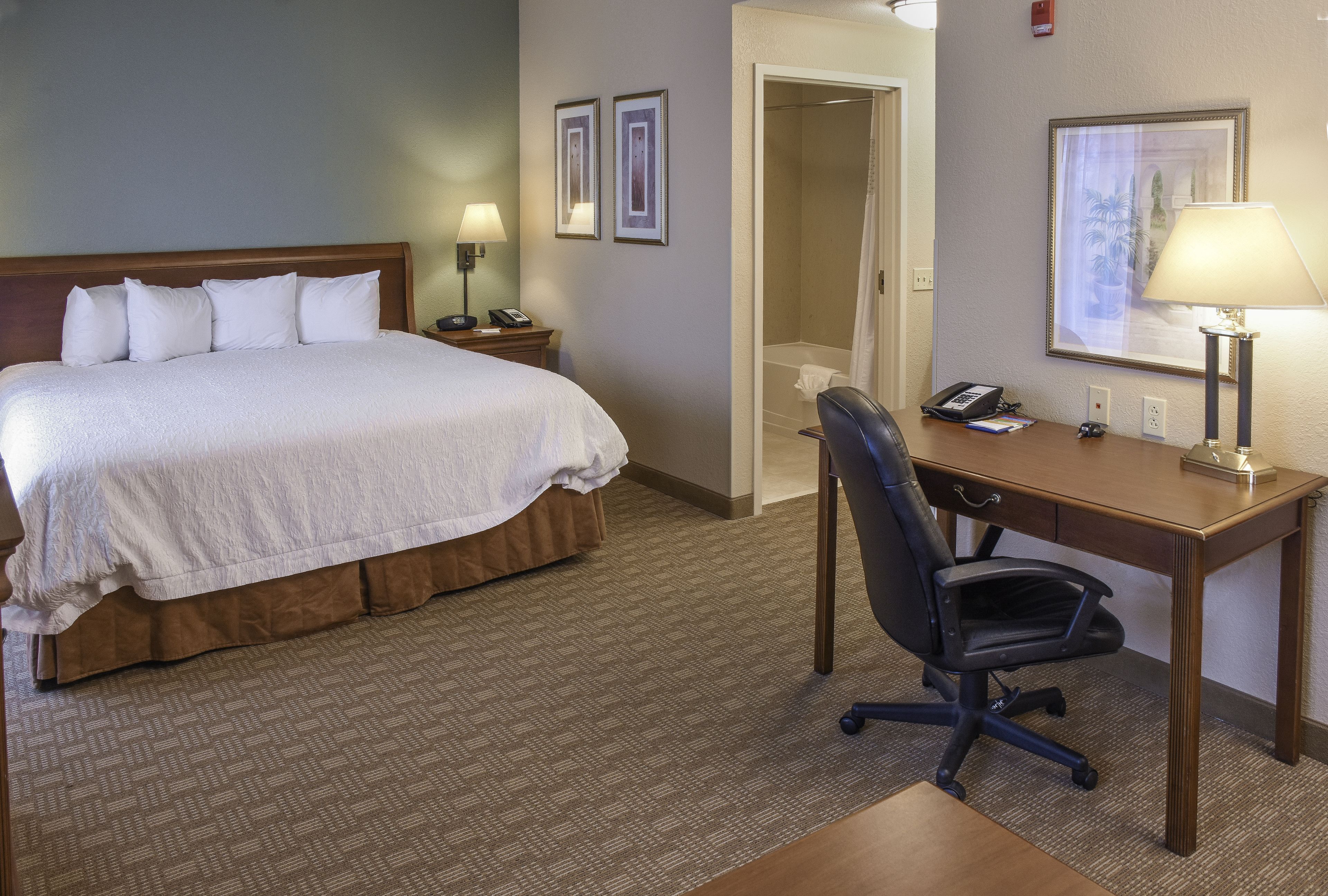 Quarto de hotel com cama, escrivaninha, cadeira e porta do banheiro.