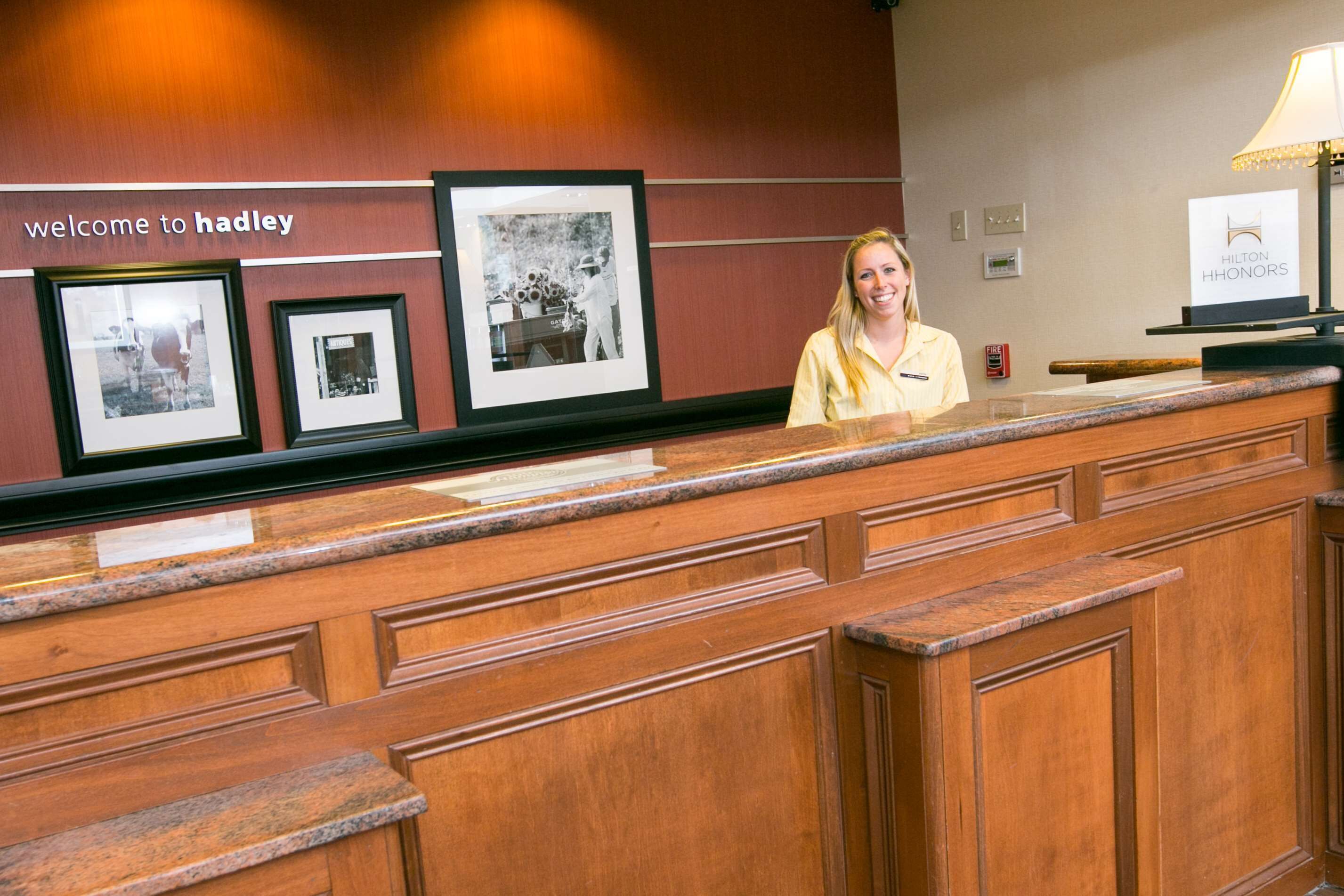 Un mostrador de recepción con fotografías enmarcadas en la pared, un cartel que dice 'welcome to hadley' y un logo de Hilton Honors.