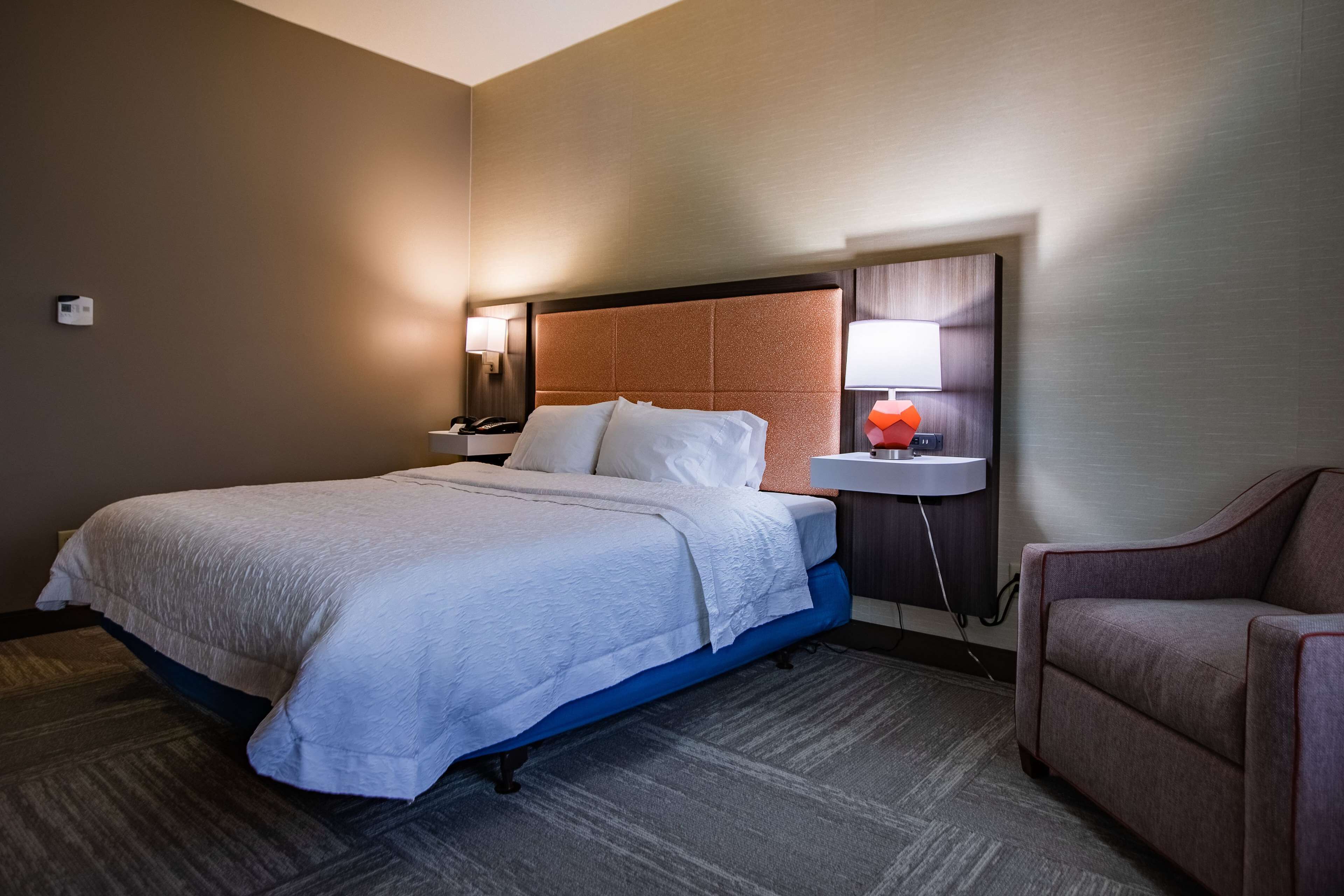 A hotel room with a large bed, a bedside table with a lamp, a small table with a red object, and a chair.