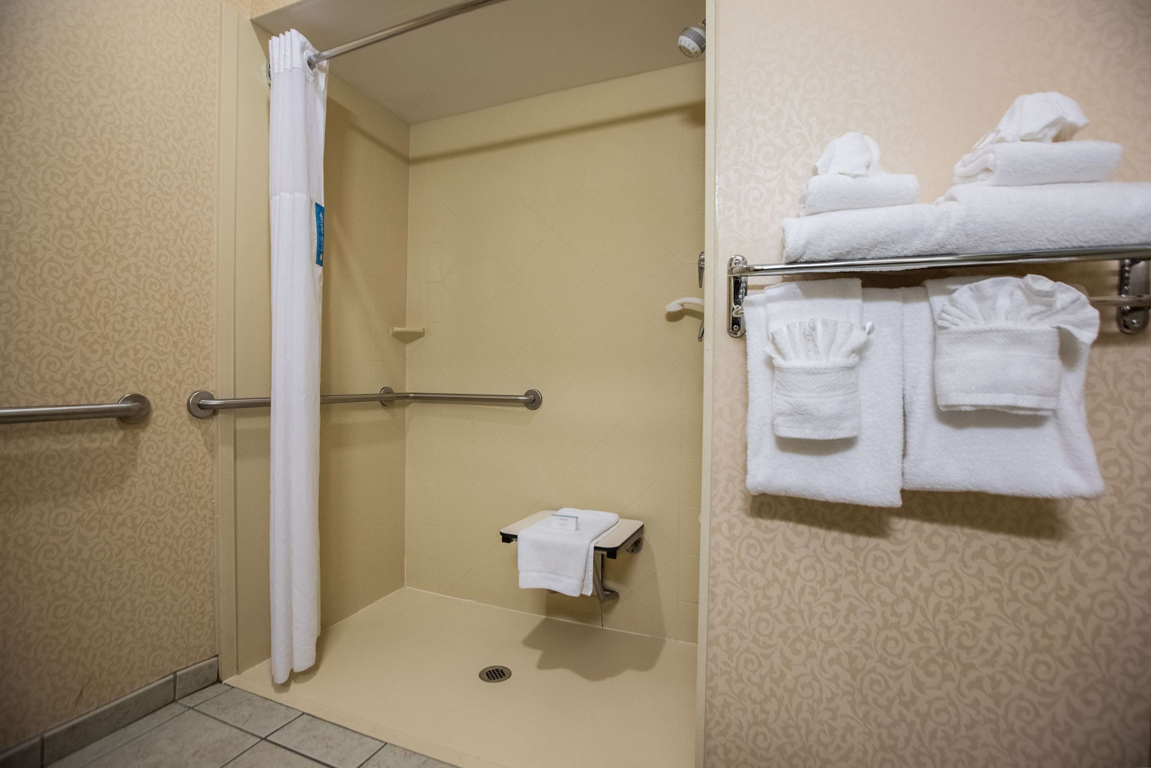 A shower area with a white shower curtain, a grab bar, a towel rack with folded towels, and a small towel dispenser.