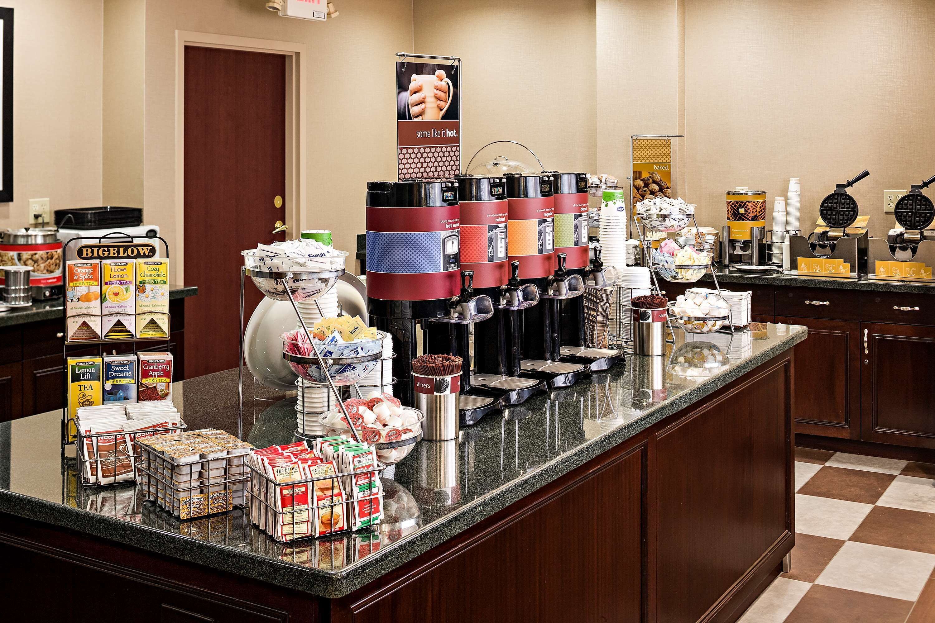 A breakfast bar with various food and drink dispensers, including a waffle maker, coffee machines, and a bowl of cereal.