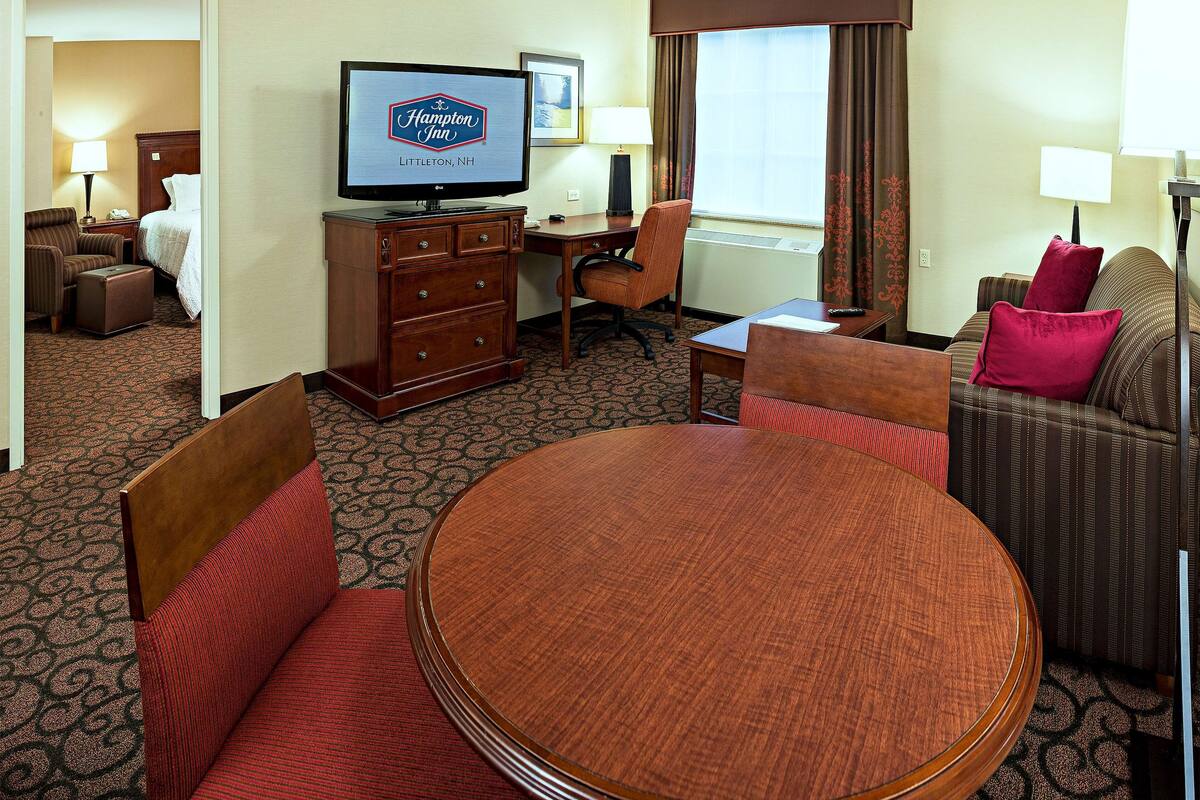 A hotel room with a round dining table, a sofa, a desk, and a television displaying the Hampton Inn logo.