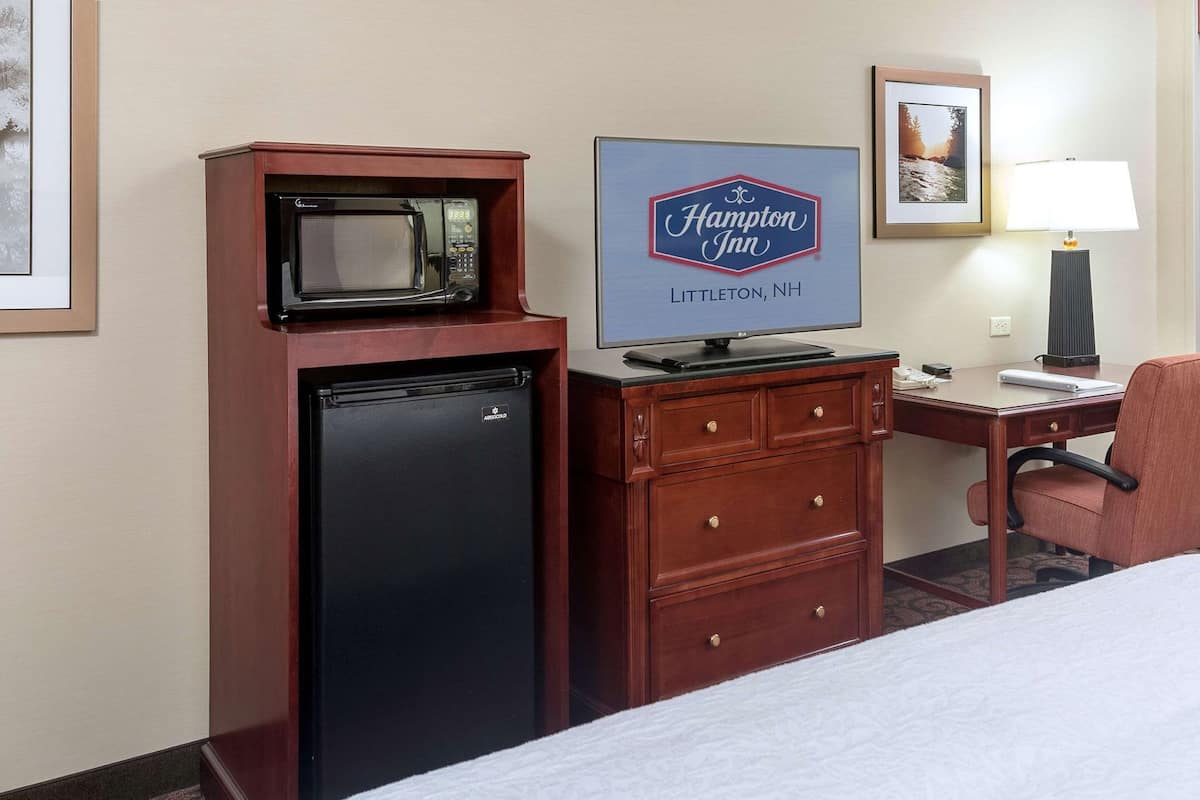 Quarto de hotel com cama, escrivaninha com computador, geladeira, micro-ondas, luminária e placa da Hampton Inn.