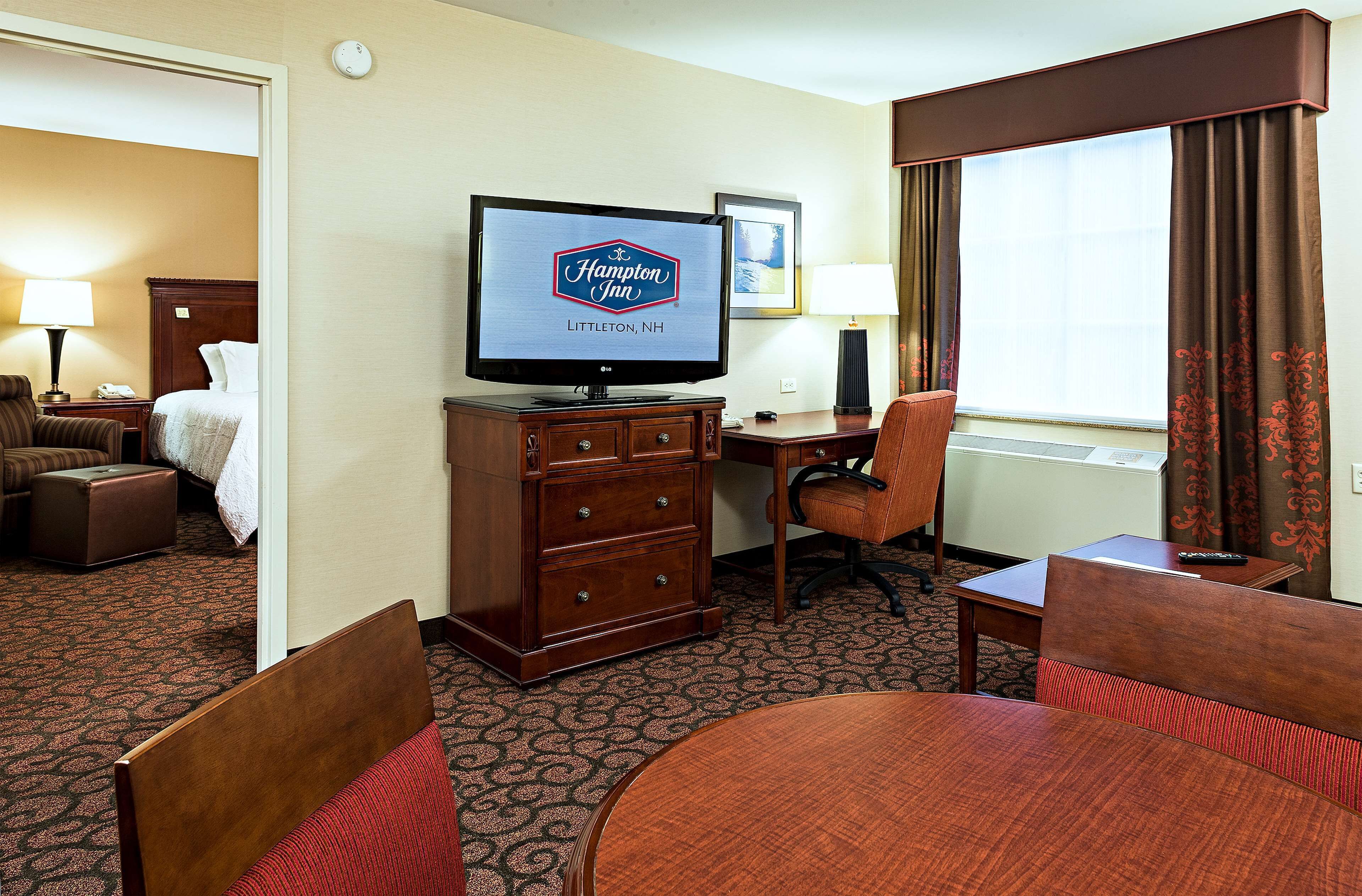 Ett hotellrum på Hampton Inn i Littleton, New Hampshire, med TV, skrivbord, stol, säng och en matplats.