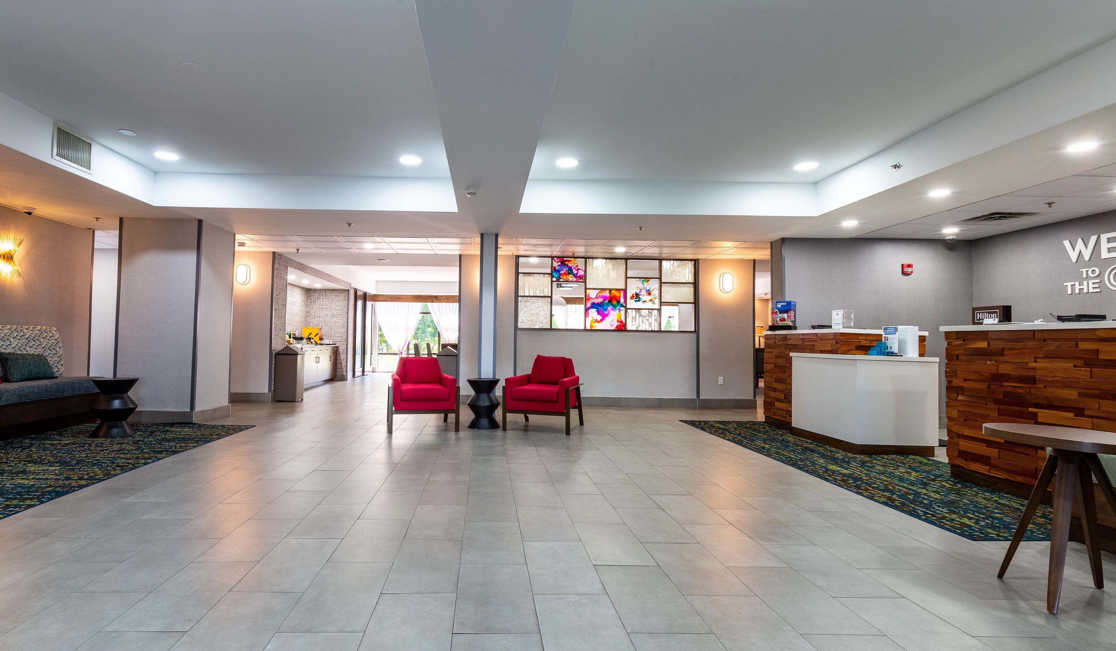 A hotel lobby with a reception desk, two red chairs, a sofa, and a large window with a colorful view.