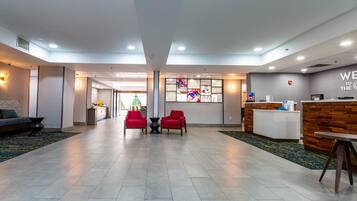 A hotel lobby with a reception desk, two red chairs, a sofa, and a large window with a colorful view.