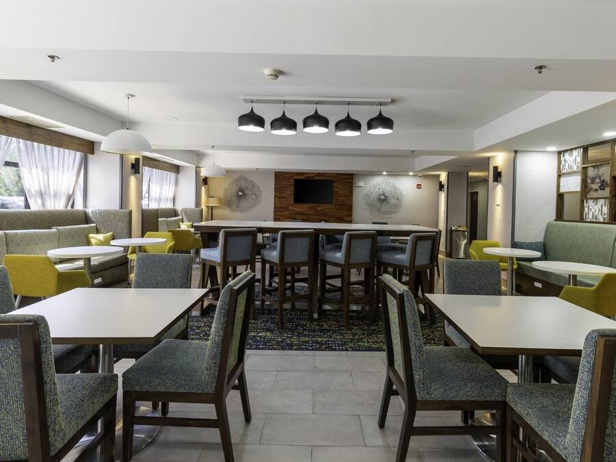 A hotel dining area with multiple tables and chairs, a central wall with a decorative panel, and a ceiling with pendant lights.